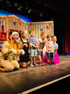 A man in a dog costume, face now revealed, sits on the stage with paws up, alongside him kneel and stand 4 young children and a mum on the end 