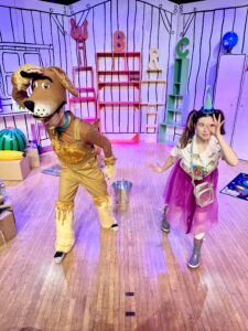 Someone in a dog costume, and a woman in a pink skirt and party hat are on a brightly lit, colourful stage