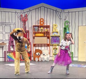 Two people are on a brightly lit stage, someone in a dog costume on the left and a woman in a pink skirt and party hat on the right