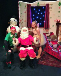 The North Pole Team (Santa in red suit and white beard flanked by an elf and a Christmas fairy) on stage in Santa In Love 