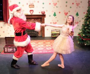 Santa in red suit, hat and white beard dances on stage with a Christmas fairy on the festive set of Santa In Love 