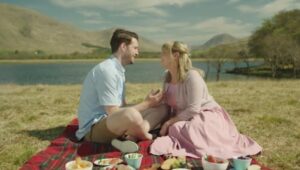 Louise and her male co-star sit on a picnic blanket on grass in front of a lake and mountains, it is a sunny day and they gaze at each other