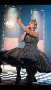 Louise, sitting on a stool, wearing a 50s-stle leopard print dress with net skirt, does her make up using a handheld mirror. Well lit room with chandelier, possibly on stage.