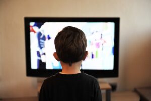 Photo of a young boy watching TV, we can only see the back of his head and the screen illuminated in front of him